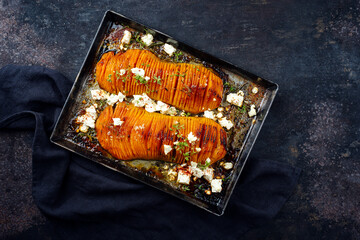 Traditional fried hasselback butternut squash pumpkin roast with herbs and feta sheep cheese served as top view in a rustic metal tray on an old black board with copy space