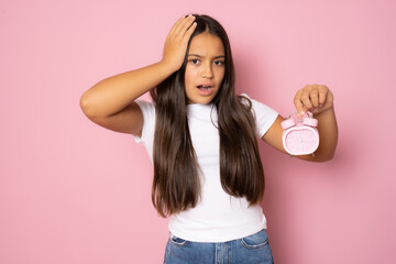 Beautiful worried girl wearing casual white t-shirt showing alarm clock standing isolated over pink background.