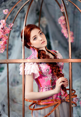 Beautiful red haired girl dressed in pink corset standing in big golden cage decorated with flowers