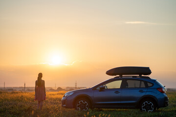 Plakat Dark silhouette of lonely woman relaxing near her car on grassy meadow enjoying view of colorful sunrise. Young female driver resting during road trip beside SUV vehicle.