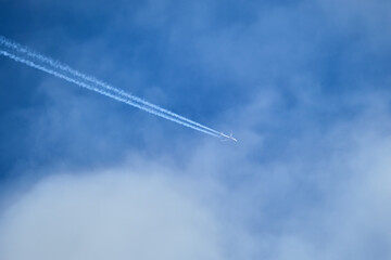 Distant passenger jet plane flying on high altitude through white clouds on blue sky leaving white smoke trace of contrail behind. Air transportation concept