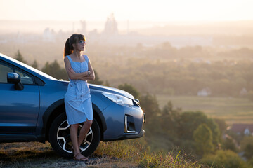 Happy young woman driver in blue dress enjoying warm summer evening standing beside her car. Travelling and vacation concept