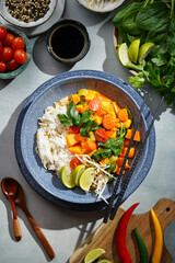 A blue bowl with red thai vegetable curry and basmati rice, fresh lime, cilantro, spinach, chilli peppers and cherry tomatoes, top view