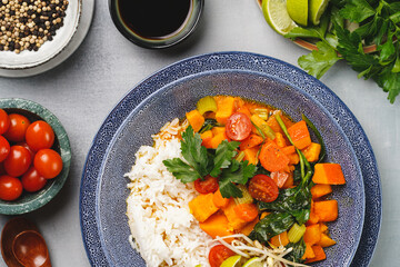A blue bowl with red thai vegetable curry and basmati rice, fresh lime, cilantro, spinach, chilli peppers and cherry tomatoes, top view