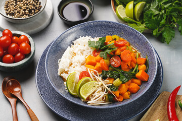 A blue bowl with red thai vegetable curry and basmati rice, fresh lime, cilantro, spinach, chilli peppers and cherry tomatoes