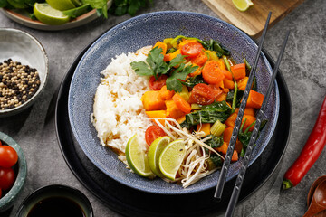 A blue bowl with red thai vegetable curry and basmati rice, fresh lime, cilantro, spinach, chilli peppers and cherry tomatoes