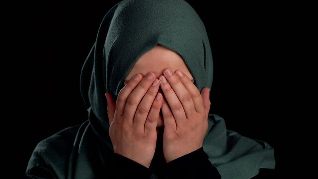Close up of a sad muslim girl's face covering her face with her hands. Isolated on a black background.