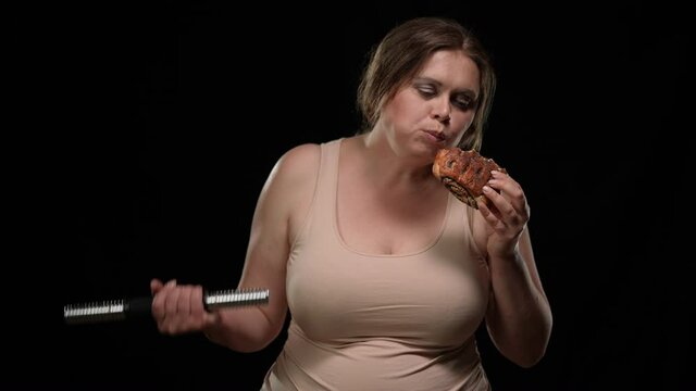 Portrait Of Obese Caucasian Woman Enjoying Taste Of Unhealthy Sweet Bun Lifting Dumbbell At Black Background. Overweight Lady Eating Delicious Junk Food Exercising