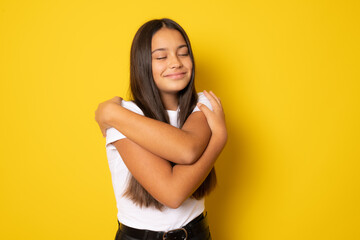 Brunette hispanic girl smiling cheerful hugging herself standing isolated over yellow background.