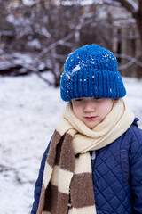Sad boy wearing bright blue knitted hat, blue jacket,  striped scarf outdoors on beautiful winter snowy day.