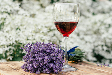 half-empty glass of red wine and lavender bouquet tied with blue ribbon on wooden brown table in sunny day at sunset in garden against organic white flower bush background. soft focus - Powered by Adobe