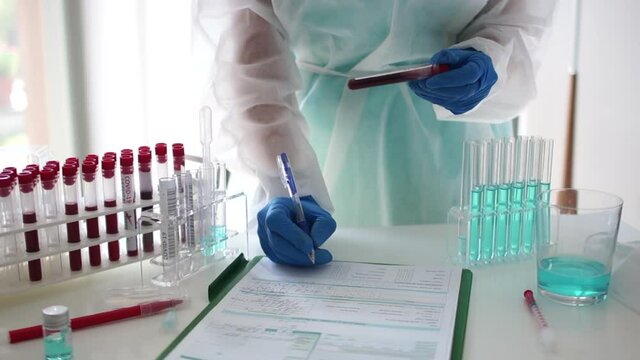 Cloce up of rack with test tubes with blood samples. Covid-19 test. Unrecognizable laboratory assistant wearing a protective suit and gloves records the results of a coronavirus test