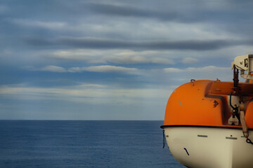 Lifeboat hanging above the decks onboard a cruise ship. Orange and white raft for use in a nautical emergency. With view of the sea with a moody sky. With space for text. - 477378448