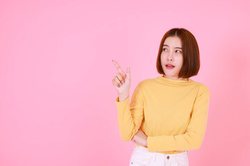 Portrait studio cutout shot of Asian young pretty short hair female model in yellow long sleeve shirt stand smile to look at the camera and pointing finger on pink background.