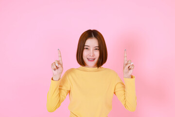 Portrait studio cutout shot of Asian young pretty short hair female model in yellow long sleeve shirt stand smile to look at the camera and pointing finger on pink background.