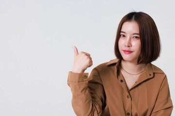 Studio cutout shot of Asian young pretty short hair female model in long brown coat jacket stand smile crossed arm hold thumb up pointing presenting product on blank copy space on white background