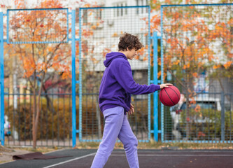 Cute teenager in violet hoodie playing basketball. Young boy with ball learning dribble and shooting on the city court. Hobby for kids, active lifestyle