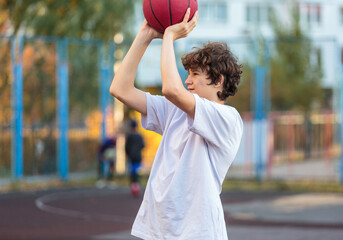 Cute teenager in white t shirt playing basketball outside. Young boy with ball learning dribble and shooting on the city court. Hobby for kids, active lifestyle