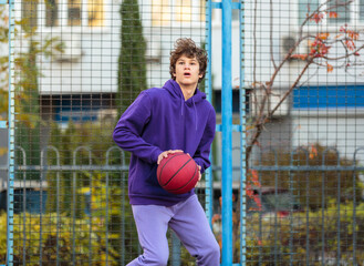 Cute teenager in violet hoodie playing basketball. Young boy with ball learning dribble and shooting on the city court. Hobby for kids, active lifestyle