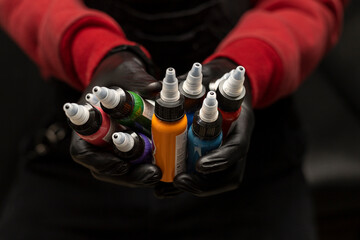 Fototapeta Male tattoo artist holding bottles of tattoo ink of various colors. Selective focus on the jars, black background, wearing red sweater with black gloves. Body art concept obraz