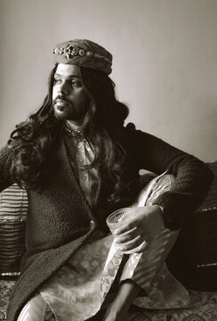 Grayscale photo of Indian man with long hair sitting on floor holding a drink