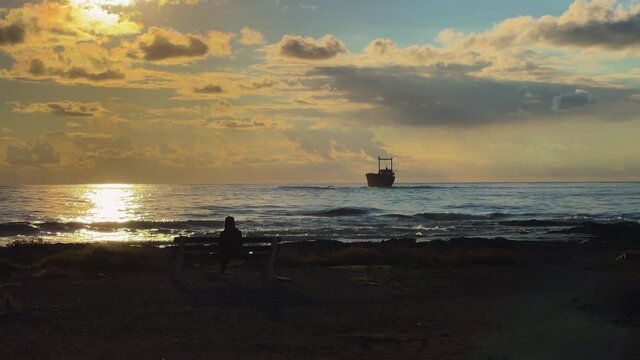 Woman sits on the shore of Cyprus during sunset. She looks out toward a shipwreck, her figure is silhouetted. Dimitrios II Shipwreck in the background.