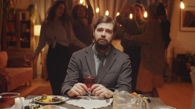 Horizontal medium portrait of sad young Caucasian man with beard on face sitting at dining table, his friends dancing behind him