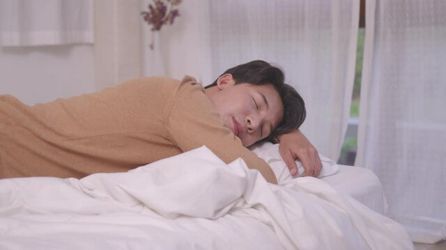 Asian man sleeping well in a comfy warm fresh bed on a soft pillow white linen orthopedic mattress, a calm serene male relaxing lying asleep in the morning.
