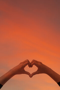 A woman raises her hand above her head to make a heart symbol on the background of the bright morning sky because the heart symbol signifies love, friendship and kindness to one another.