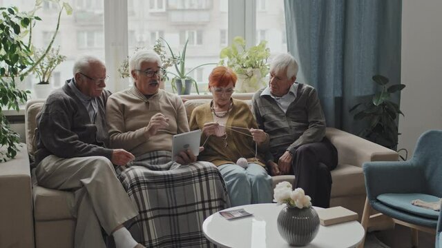 Four Caucasian seniors sitting on sofa in nursing home spending leisure time together having conversation. Senior man holding digital tablet in hands