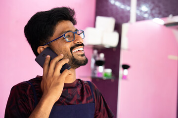 happy barber busy talking on mobile phone call at saloon - Concept of successful business owner, positive emotion and happy custome feedback showing with copy space