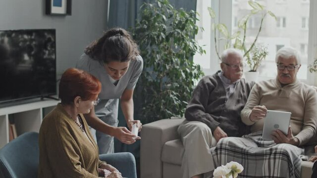 Young female nurse giving pill organizer box to senior woman knitting in chair while three senior men talking on sofa nearby, spending leisure time together at nursing home