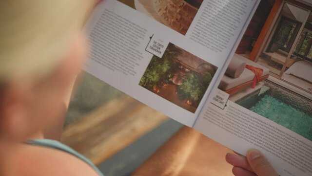 Behind blond woman reading and holding travel magazine while laying on lounge bed