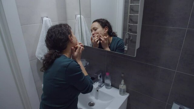 Beautiful black woman checking facial skin, removing pimple from her face while looking at mirror in bathroom home. Adult female doing morning routine, noticing zit on face during washing procedure