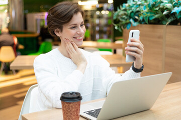 Young beautiful pretty positive brunette woman wearing white sweatshirt sitting at a shopping center at a table and working at a computer laptop, using mobile phone. Freelance and business concept
