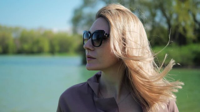portrait of blonde woman in sunglasses on the background of the lake in the summer park. Hair develops in the wind.