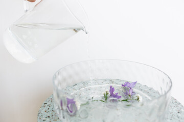 water is poured into a glass basin with purple flowers floating in the water. water pours from a glass jug.