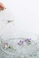 water is poured into a glass basin with purple flowers floating in the water. water pours from a glass jug.