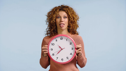 young woman with red wavy hair sticking out tongue while holding round clock isolated on blue