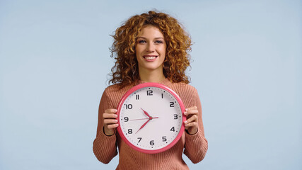 happy woman with red wavy hair holding wall clock isolated on blue