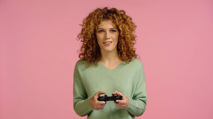 KYIV, UKRAINE - DECEMBER 22, 2021: smiling redhead woman gaming with joystick isolated on pink