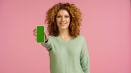 cheerful redhead woman looking at camera while showing smartphone with green screen isolated on pink