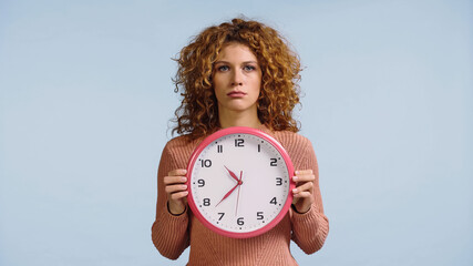 upset woman with round clock looking at camera isolated on blue