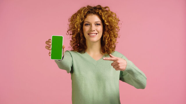 blurred cheerful woman pointing at mobile phone with green screen isolated on pink
