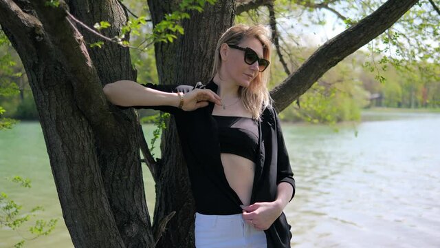 A beautiful young woman in sunglasses in a black top and an unbuttoned shirt poses in nature near a lake standing under a tree