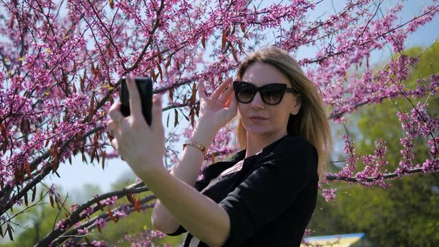A pretty young blonde woman in sunglasses takes a selfie on her phone on a beautiful background of cherry blossoms