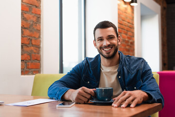 cheerful man looking at camera near cup of coffee and smartphone with blank screen in cafe
