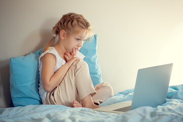 A cute girl sitting on the bed is watching something on a laptop. The concept of education.