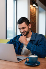 smiling man looking at laptop near blurred coffee cup in cafe