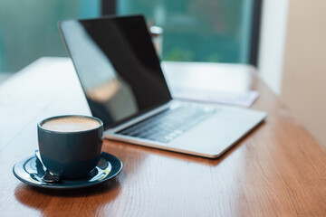 selective focus of cup with cappuccino near blurred laptop with blank screen on table in cafe